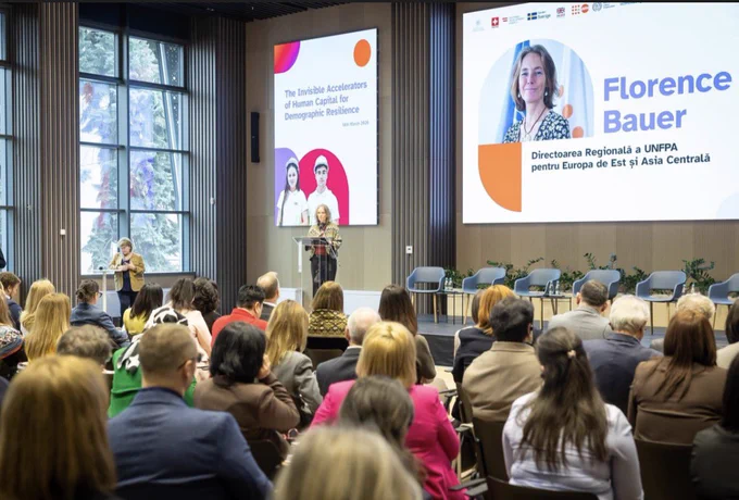 A conference hall where Florence Bauer, UNFPA Regional Director for Eastern Europe and Central Asia, stands at a podium giving a presentation. A large screen to her right displays her name and portrait, while a screen to her left shows the presentation title: "The Invisible Accelerators of Human Capital for Demographic Resilience," dated March 18, 2026. An audience is seated in the foreground, facing the stage in a modern room with large windows and wooden wall paneling.