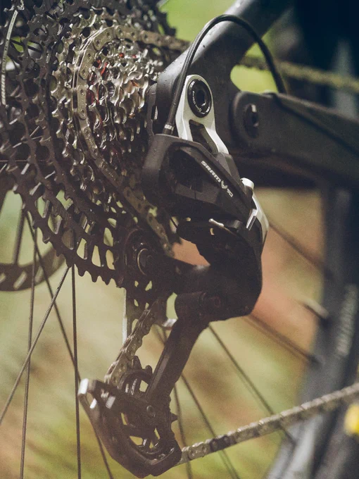  Close-up image of a mountain bike rear derailleur and gear cassette. The derailleur is SRAM Eagle Mechanical Transmission in an outdoor setting accompanied with a blurry image showing a mountain bike rider at speed.