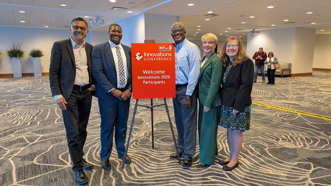 A group of five people stand smiling next to a red sign that reads, "Welcome Innovations 2026 Participants" at the Innovations Conference. The setting is a modern indoor conference space.