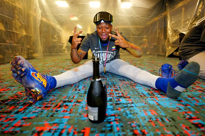 Ronald Acuña Jr. sits on the floor of a clubhouse following Team Venezuela’s World Baseball Classic Final win. He wears white pants, a white cap, a gray shirt, goggles and a gold medal. He has one shoe on and one off. He is making a peace sign motion with both of his hands. His phone leans on a bottle of champagne in front of him.