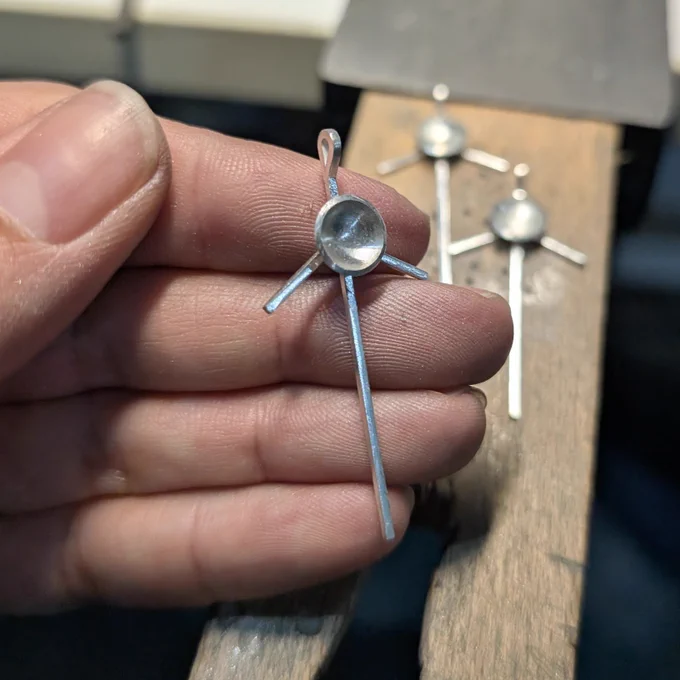 Hand holding a sterling silver Voyager spacecraft pendant, two more pendants on the workbench behind it