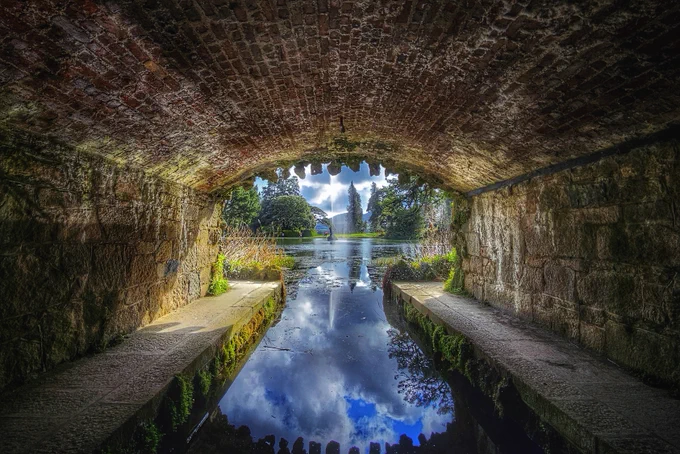 A low-angle shot taken from inside a dark, rustic brick and stone tunnel, looking out toward a bright, sunlit garden lake. The curved brick ceiling and stone side walls create a natural frame. In the centre, a calm lake perfectly reflects a blue sky with fluffy white clouds and a tall water fountain spray. Lush green trees and a distant hillside line the horizon under a bright, ethereal light. The foreground water is dark and still, acting as a mirror.
