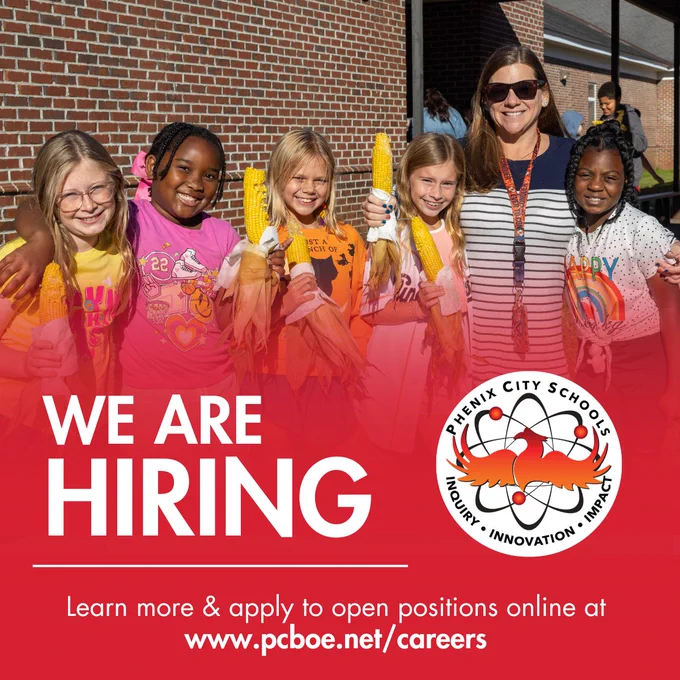 A promotional image for Phenix City Schools hiring featuring a smiling female teacher with her arm around five diverse female elementary school students. The group is outside a brick school building on a sunny day, holding ears of fresh corn. The bottom third of the image is a solid red field with large white text stating "WE ARE HIRING," the district logo, and a line of text "Learn more & apply to open positions online at www.pcboe.net/careers."