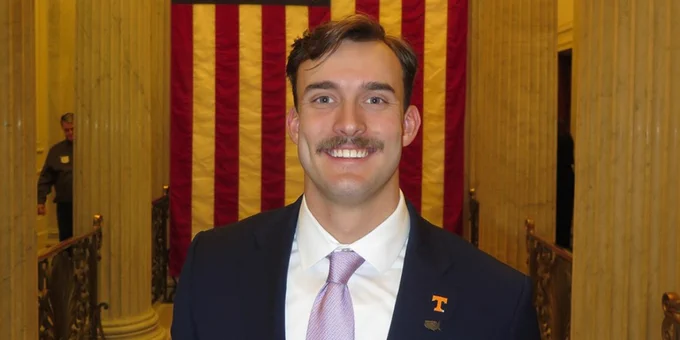 Wildlife and fisheries science student wearing a suit and tie stands in a grand hallway in the US Capitol Building. An American flag is prominently displayed in the background.