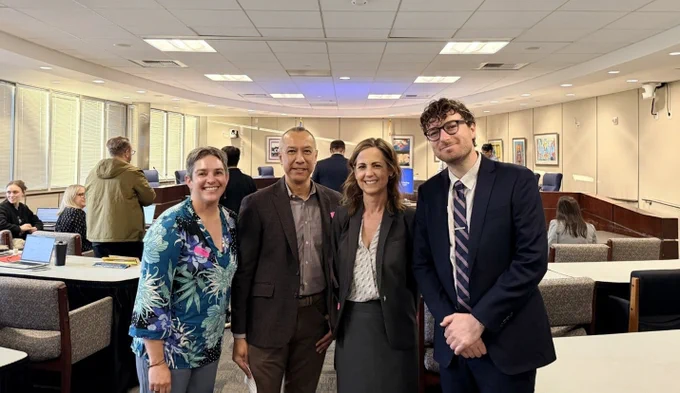 Four people stand in a well-lit conference room smiling at the camera. Left to right: Jenny Scala, Ben Kalina, Christine Olmstead, Leib Sutcher. There are tables and chairs, with other individuals working in the background. Framed pictures are on the walls. 