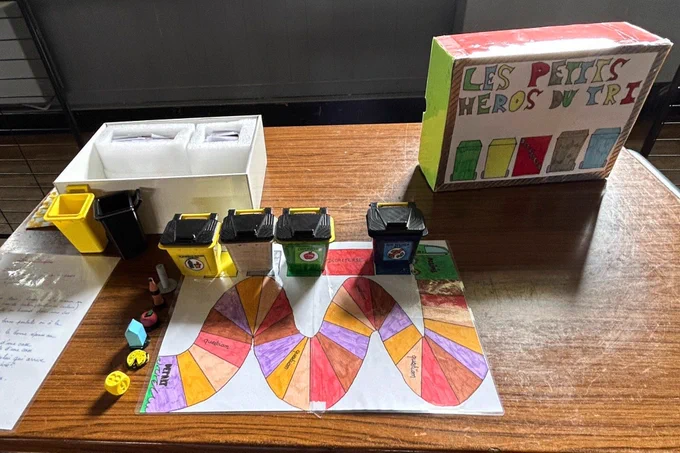 Jeu pédagogique sur le recyclage installé sur une table en bois. On voit plusieurs petites poubelles miniatures de couleurs différentes (jaune, vert, bleu, noir), un plateau, des pions et une boîte intitulée "Les petits héros du tri". 