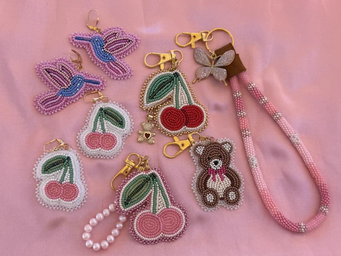 Assortment of beadwork, flat stitched cherry bag charms/keychains (pink and red), teddy bear bag charm/keychains, humming bird earrings, and pink cherry earrings. Laying on pink satin.