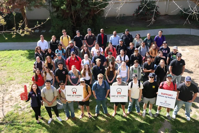 A group of people are gathered outdoors on a grassy area. Some hold signs reading "Day of Giving" and "Support Future Ag Leaders", referencing California State University, Fresno. The setting is sunny with trees in the background.
