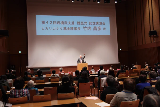 ステージ中央の演台に立ち、講演を行う竹内先生の写真です。
左腕をあげ、人差し指を上にあげています。
多くのボランティアの方が座って講演を聞いています。