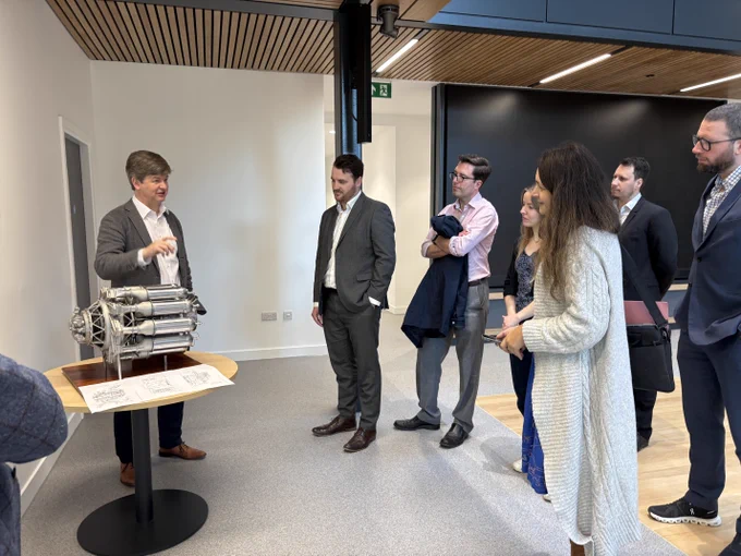 A group of people in formal attire are gathered around a speaker who is pointing at a mechanical object on a table. The setting appears to be a modern office space with wooden ceiling panels and white walls. The object has an intricate design with multiple cylindrical components. There are papers laid out next to it. The group is attentively listening to the speaker.