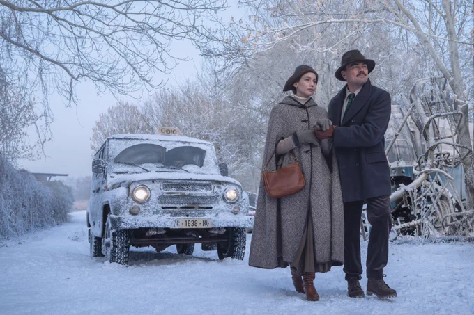 A first look at Martin Scorsese's new film titled, "What Happens at Night." The photo features a rural setting, the ground and nearby bushes are covered in snow. In the foreground, Jennifer Lawrence and Leonardo DiCaprio are walking hand in hand towards a destination off screen. They are dressed in period attire. They have concerned looks on their faces. A taxi idles behind them, covered in snow. 