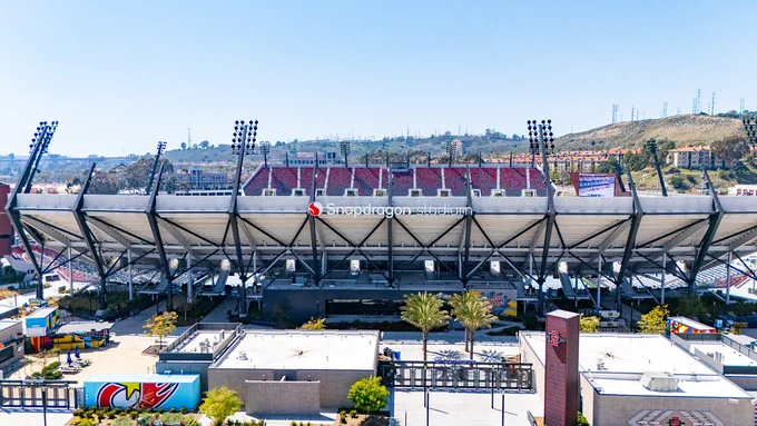 A large stadium with prominent lighting structures, named Snapdragon Stadium, is pictured. The seating area is visible above the main structure. There are some palm trees and small buildings in the foreground, and a hillside with various structures in the background.