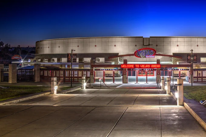 Exterior view of Viejas Arena at night, with illuminated signs and entrance. The paved walkway leads up to the arena, and the sky is deep blue.
