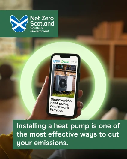 Image of someone holding a phone with a photograph on the phone screen of a heat pump  with a caption under that reads "Discover if a hear pump could work for you". Text on image underneath reads "Installing a heat pump is one of the most effective ways to cut your emissions".