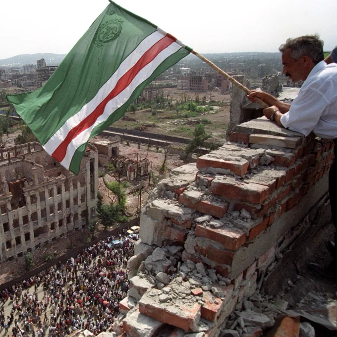 On the righthand side of this image, an older man waves the flag of the Chechen Republic of Ichkeria — colored green, white, and red — over a crowd below as he leans on rubble of a brick wall formerly constituting the rooftop of a building. Beige-colored, destroyed buildings can be seen in the background, and a crowd of what appears to be the city’s inhabitants flows through the street below.  