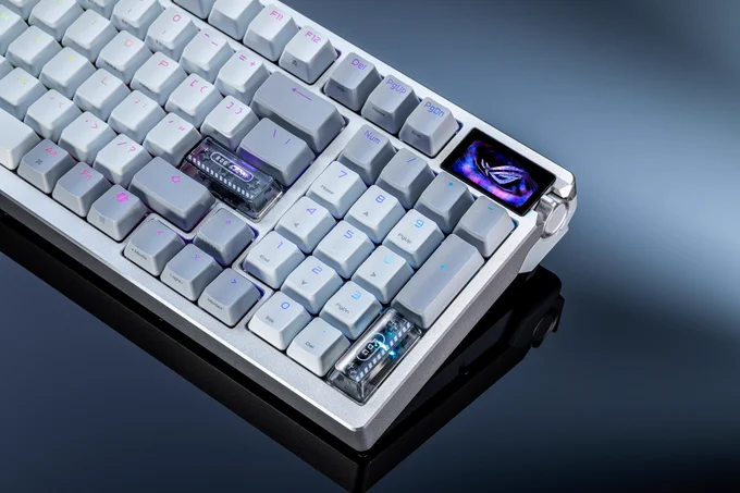 Clavier gaming compact ROG Azoth 96 HE en version blanche, avec touches rétroéclairées RGB, switches visibles et écran OLED affichant le logo ROG, posé sur une surface sombre avec reflet.