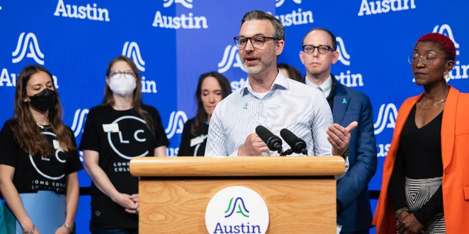 A person speaks at a podium with the Cit of Austin logo, surrounded by several individuals. The background features repeated City of Austin branding. Some people wear "Long COVID Collective" shirts.