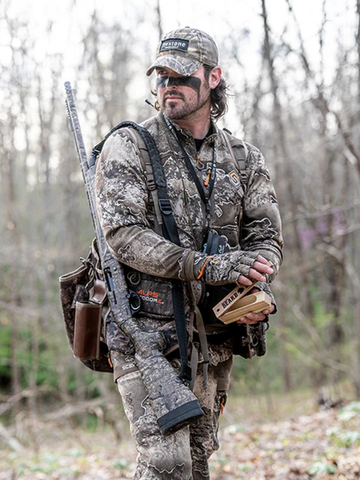 A person dressed in full camouflage gear and holding a box call stands in a wooded area. They are carrying hunting equipment, including a backpack and a shotgun. The background shows leafless trees and sparse greenery.