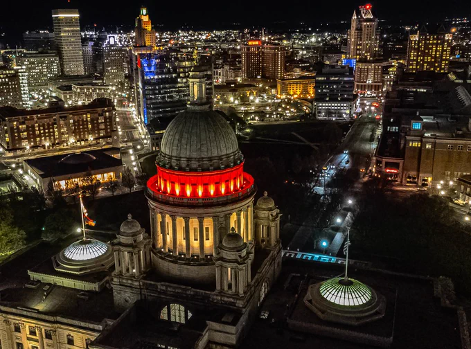 Aerial view of downtown Providence, Rhode Island at night. The Rhode Island State House is prominently lit, with its dome glowing red. The city skyline is illuminated with buildings and streetlights visible in the background.
