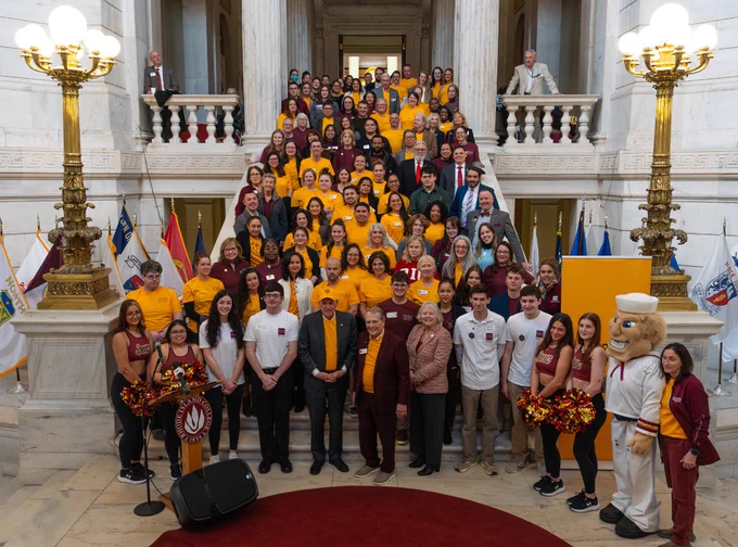 A large group of people, some in maroon or yellow shirts, are gathered on a grand staircase inside a spacious building. Flags are displayed on both sides. 
