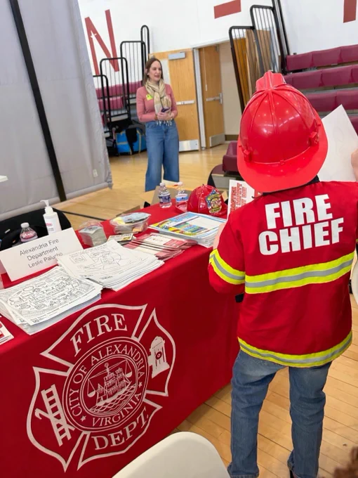 The image shows a child wearing a "Fire Chief" jacket and a firefighter helmet standing at a table with a red tablecloth that reads "City of Alexandria, Virginia Fire Dept." The table is covered with coloring pages, crayons, and water bottles. Another person is visible in the background near the bleachers, engaged in conversation.