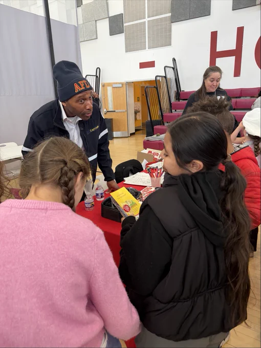 A person wearing an "AFD" beanie is engaging with a group of children around a table with pamphlets and items. The group stands in what appears to be a gymnasium with bleachers and a basketball hoop visible in the background.