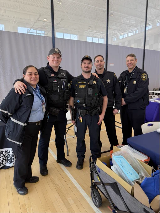 A group of five uniformed law enforcement officers stand together in a gymnasium. They are smiling and have various equipment attached to their uniforms. In front of them, there's a cart filled with supplies.