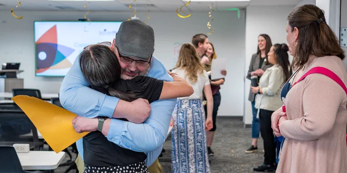 ActivateWork alumnus Fred Delehoy warmly embracing his career coach, Kelli Turner, at his cohort's graduation, while others chat and smile in the background and his wife stands by, smiling proudly.