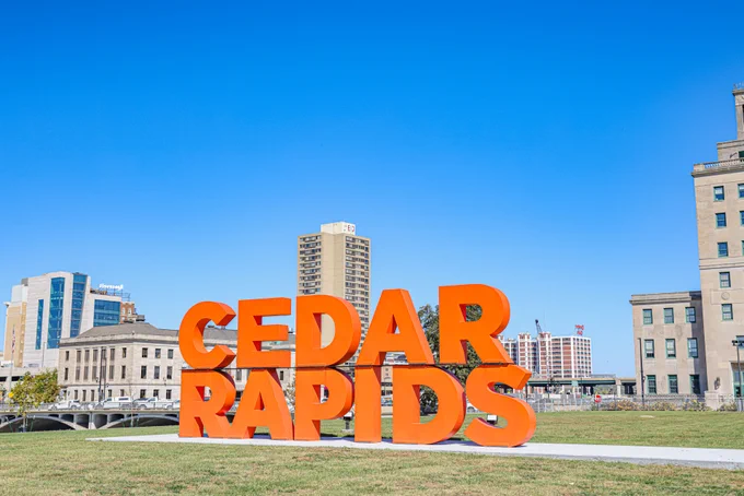 A large, vibrant orange sign displays the words "Cedar Rapids" in a grassy area. In the background, there are several buildings and a clear blue sky.