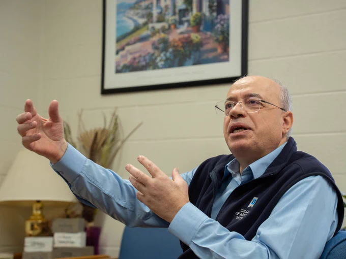 Professor Haitham Alkhateeb speaks during an interview, gesturing with his hands while seated in an office, with a framed landscape artwork and desk items in the background.