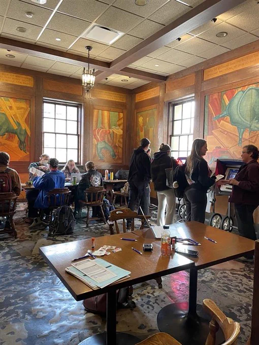 Voters gathered in a warmly lit room with large windows and colorful murals, poll workers seated at tables and others standing and conversing waiting to vote at the UW-Madison Memorial Union's Bunyon Room. 