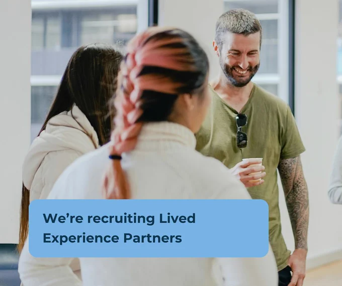 A group of three adults standing indoors in conversation. One person is holding a cup. A blue box in the foreground contains the text: ‘We’re recruiting Lived Experience Partners.’