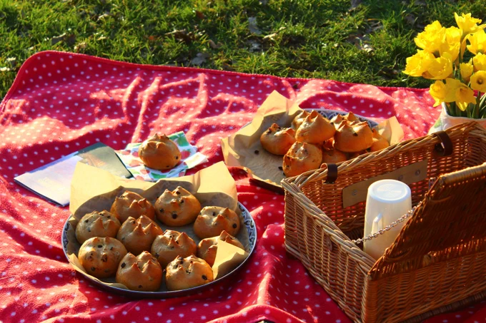 Two bowls of fruited buns, a vase of daffodils and a wicker picnic basket are laid out on a red polka dot blanket, with grass visible in the background