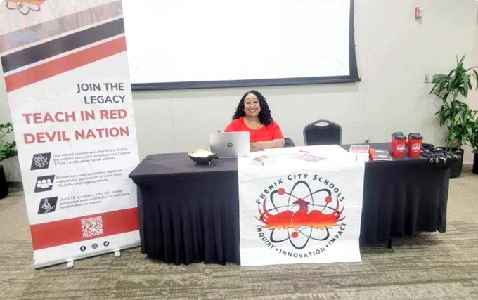 Recruitment booth for Phenix City Schools with a banner promoting teaching in the Red Devil Nation and a representative seated behind the table.
