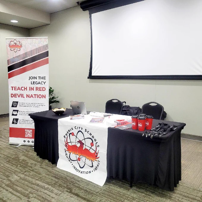 Phenix City Schools recruitment table with branded mugs, lanyards, and informational materials in a conference room.