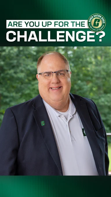 Mark Collard stands outdoors in front of blurred green foliage, wearing glasses, a white collared shirt, and a dark blazer with an All‑In‑C pin on the lapel. A green banner at the top reads “Are you up for the challenge?” alongside a circular “Niner Nation Gives — For the Love of Charlotte” badge.