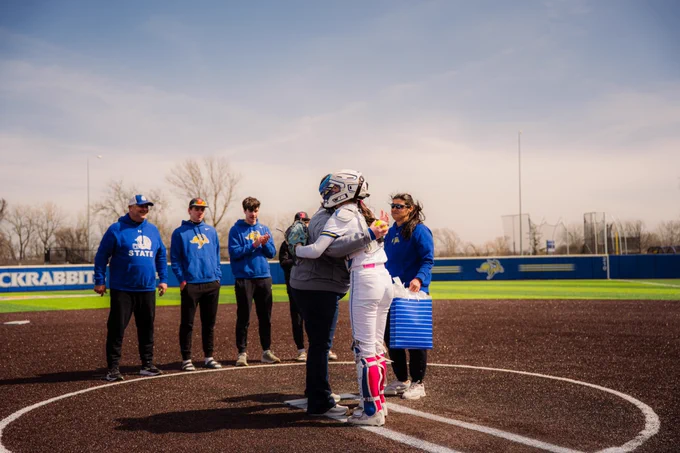 Two people hug in the circle of a softball diamond. Five other individuals look on with the sun out during a chilly day.