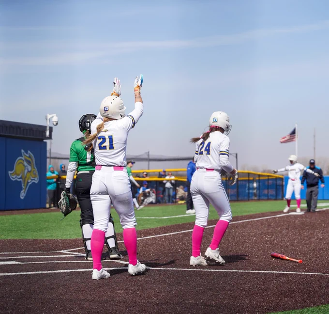 Two softball players in white uniforms with pink socks cheer for a teammate at third base on a softball diamond. 