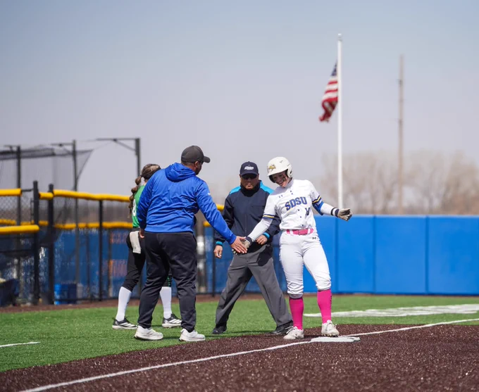 A woman in a white softball uniform with pink socks does a low five with a coach in blue and black clothing at third base on a softball diamond