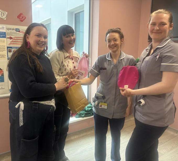 Four people are standing together in a room with pink walls. Two of them are wearing uniforms, possibly medical. They are smiling and holding items, including a paper bag and handmade pink pouches. A poster on the wall has the NHS logo.