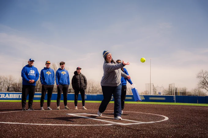A woman throws a softball towards home plate while in the circle of a softball diamond. She is watched by five individuals behind her as everybody wears layered clothing on a chilly day.