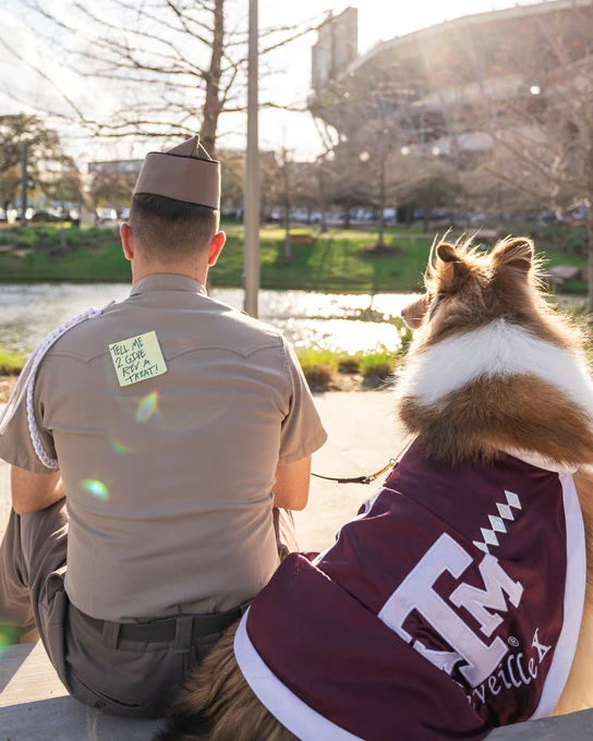 A person in uniform sits on a ledge next to Reveille, Texas A&M's mascot, overlooking a pond. They have a note on their back that reads, "Tell me 2 give Rev a treat!".