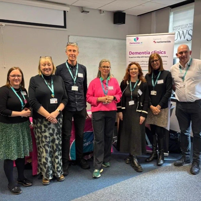 A group of seven people standing in a row indoors, all wearing name badges and smiling. Behind them is a banner displaying logos for Dementia UK. The setting appears to be a conference room or an office space.