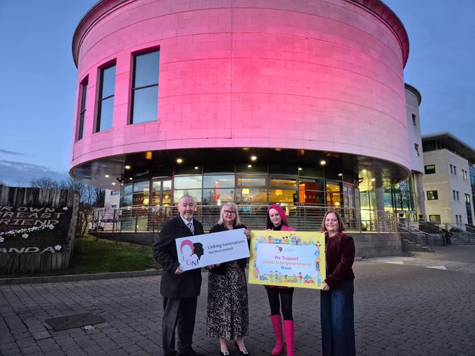 Photo shows Councillor Brian Higginson, Chair of Corporate Services, Joyce Savage and Vicki Titterington from Linking Generations NI, and Councillor Gretta Thompson, LCCC's Age Friendly Ambassador. The hold signs with the Linking Generations Logo and Intergenerational Week. All are standing in front of Lagan Valley Island which is illuminated pink.