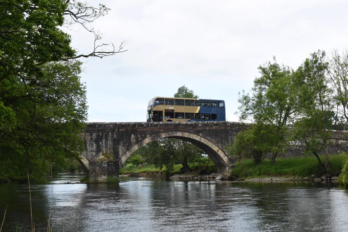 A double-decker bus driving over a stone bridge over a river, with a grass bank and trees