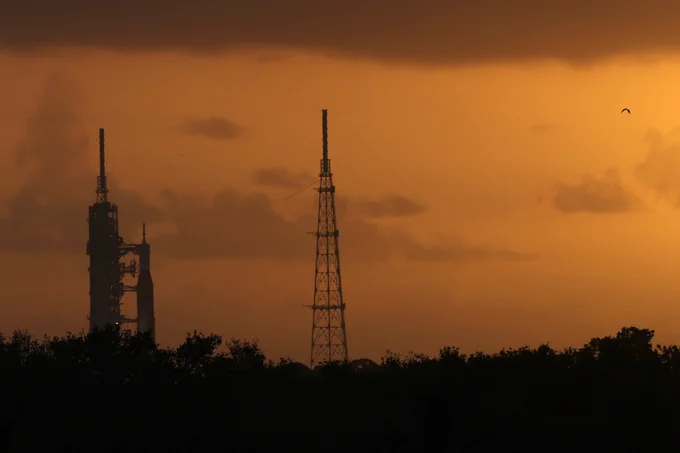 The Artemis II rocket and spacecraft can be seen at Launch Complex 39B at NASA's Kennedy Space Center during the early morning of Wednesday, April 1. The SLS (Space Launch System) rocket and Orion spacecraft are standing to the right of the trussed structure of the mobile launcher. A lightning tower can be seen in the center of the image. The sky is an orange haze in the background. Credit: NASA/Michael DeMocker 