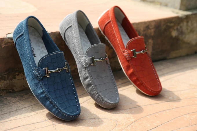 Three loafers in blue, gray, and red with textured patterns and metal buckles, displayed on a stone surface.