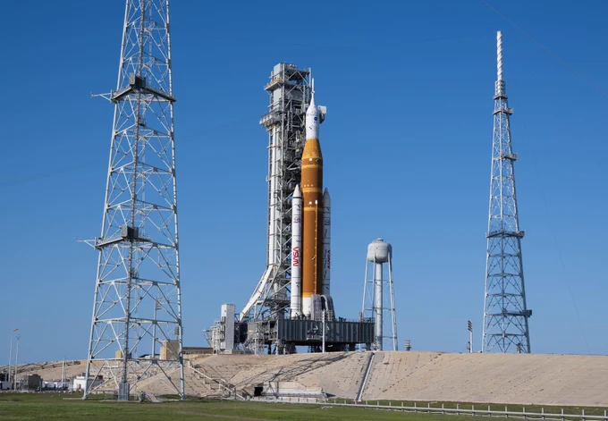 Image shows NASA's Artemis II rocket with the core stage in orange and two white boosters on each side with the NASA insignia and the America 250 insignia. On top of the orange section is the Orion spacecraft, which will carry four astronauts around the Moon. The rocket stands vertical on a silver mobile launcher 1 at Launch Complex 39B at NASA's Kennedy Space Center in Florida. Photo credit: NASA/Aubrey Gemignani