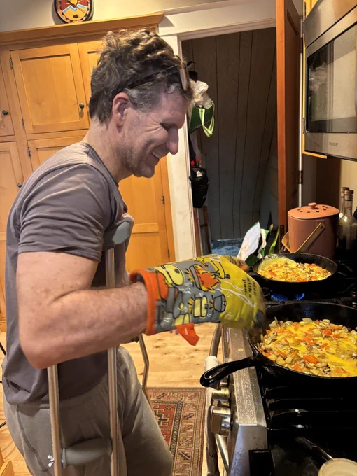 A man smiles and uses crutches while cooking two skillets of food on a stovetop. He wears an oven mitt and has sunglasses on his head, in a kitchen with wooden cabinets.