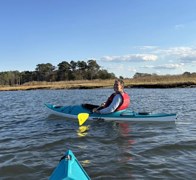 Eye-level view of a man in a kayak, wearing a red life jacket. Another kayak is in the foreground, with trees and sky in the distance. The brand \"eddyline\" is visible on the kayak.